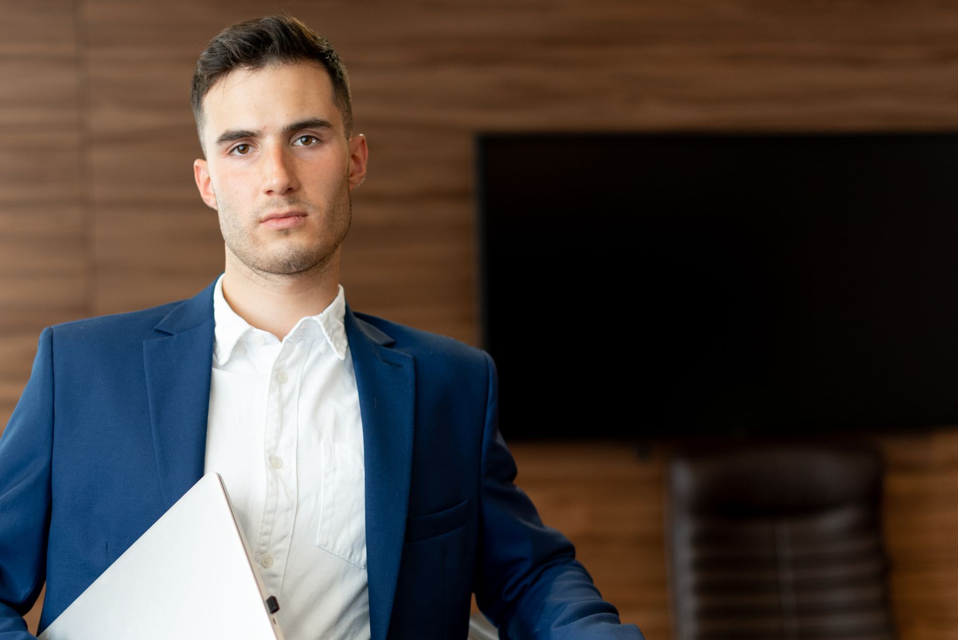 Confident young businessman in a blue suit, holding a laptop in an office setting.