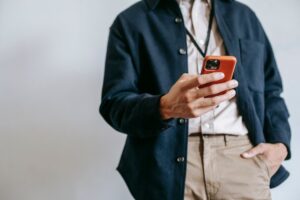 Low angle of crop unrecognizable male manager in trendy outfit and name badge messaging on mobile phone while standing near white wall with hand in pocket